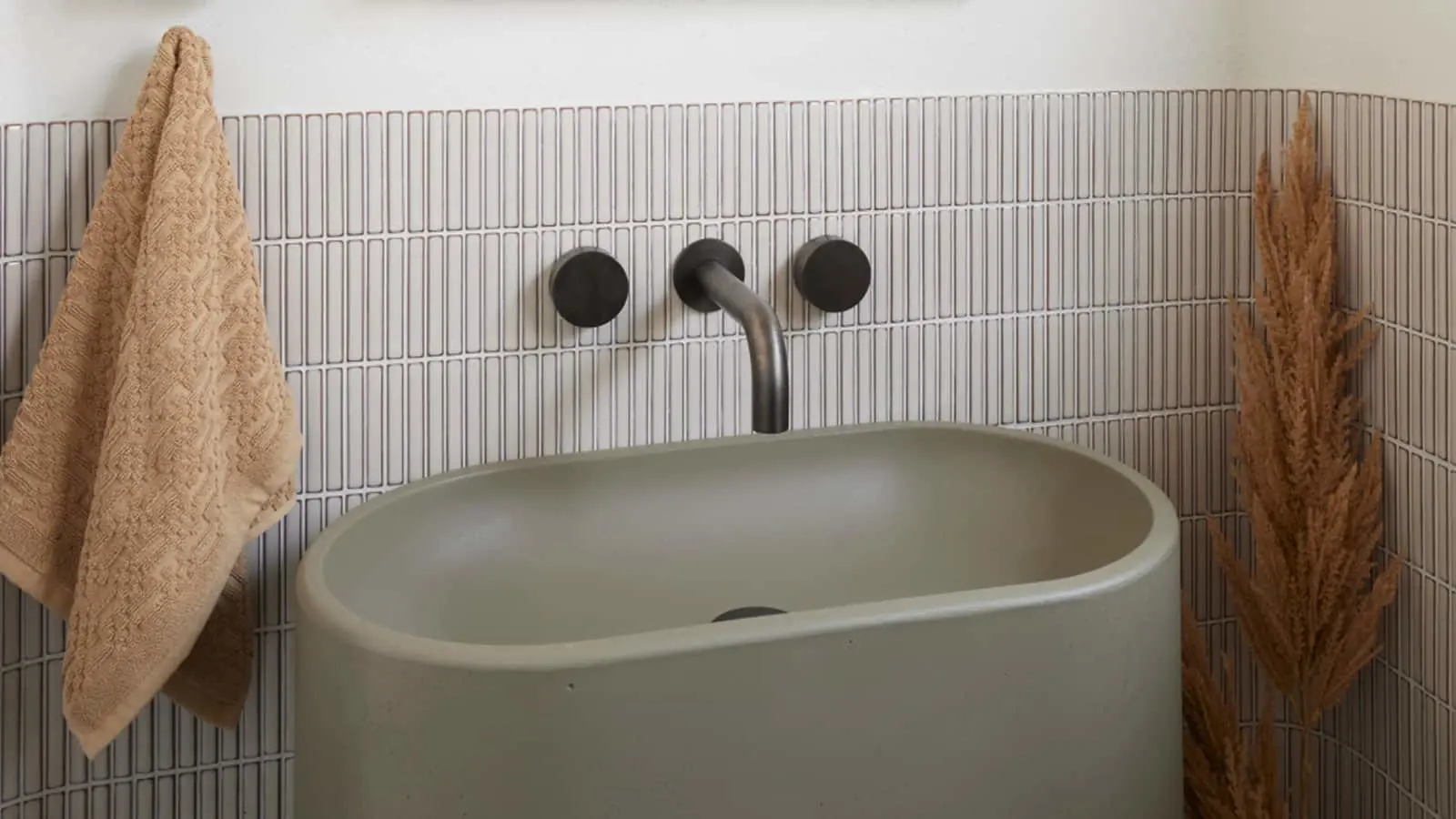 Modern bathroom sink with matte green basin, black wall-mounted faucet, beige towel, and dried pampas grass decoration.
