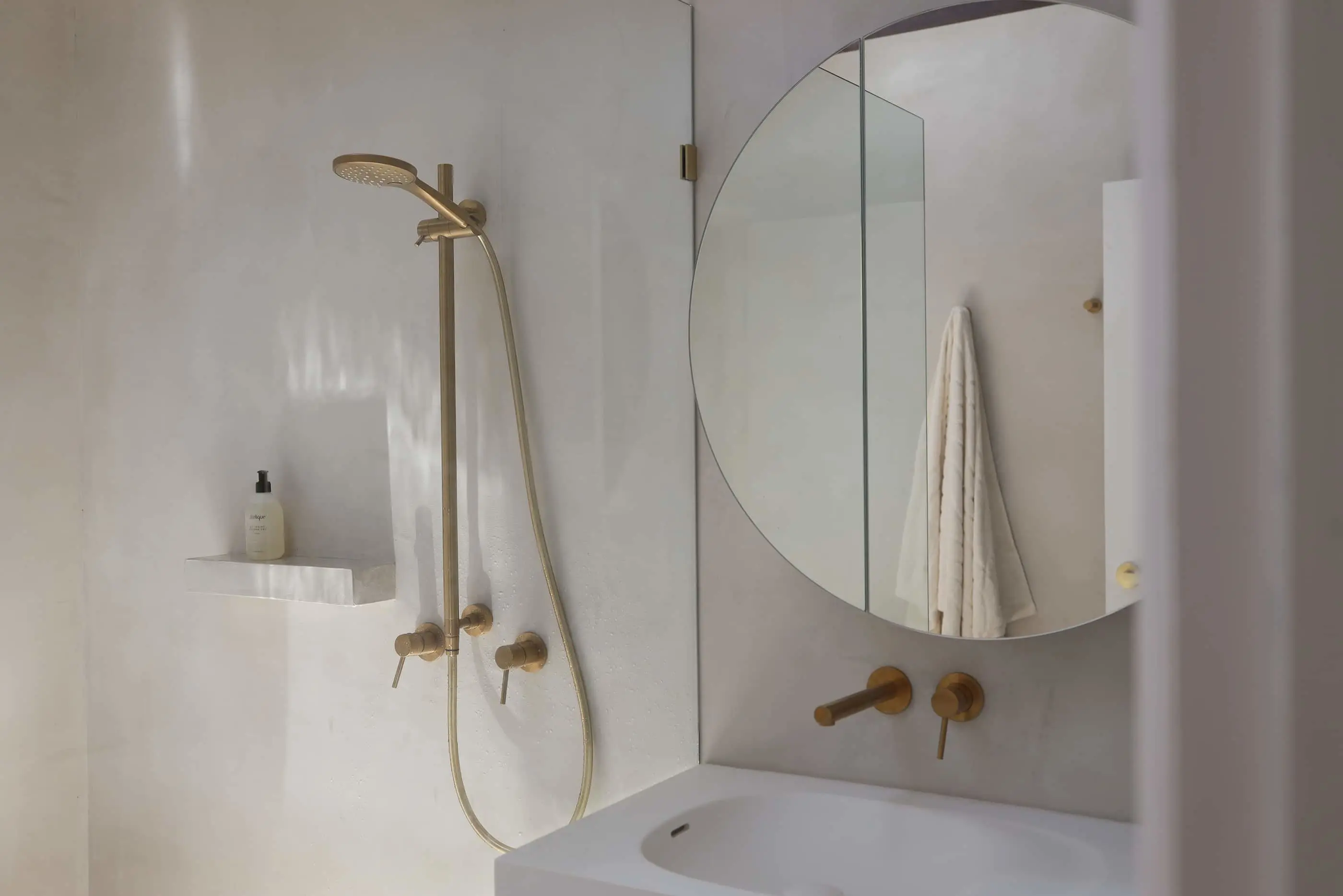 Modern white bathroom with brass fixtures, round mirror, white sink, and shower with built-in shelf for toiletries.
