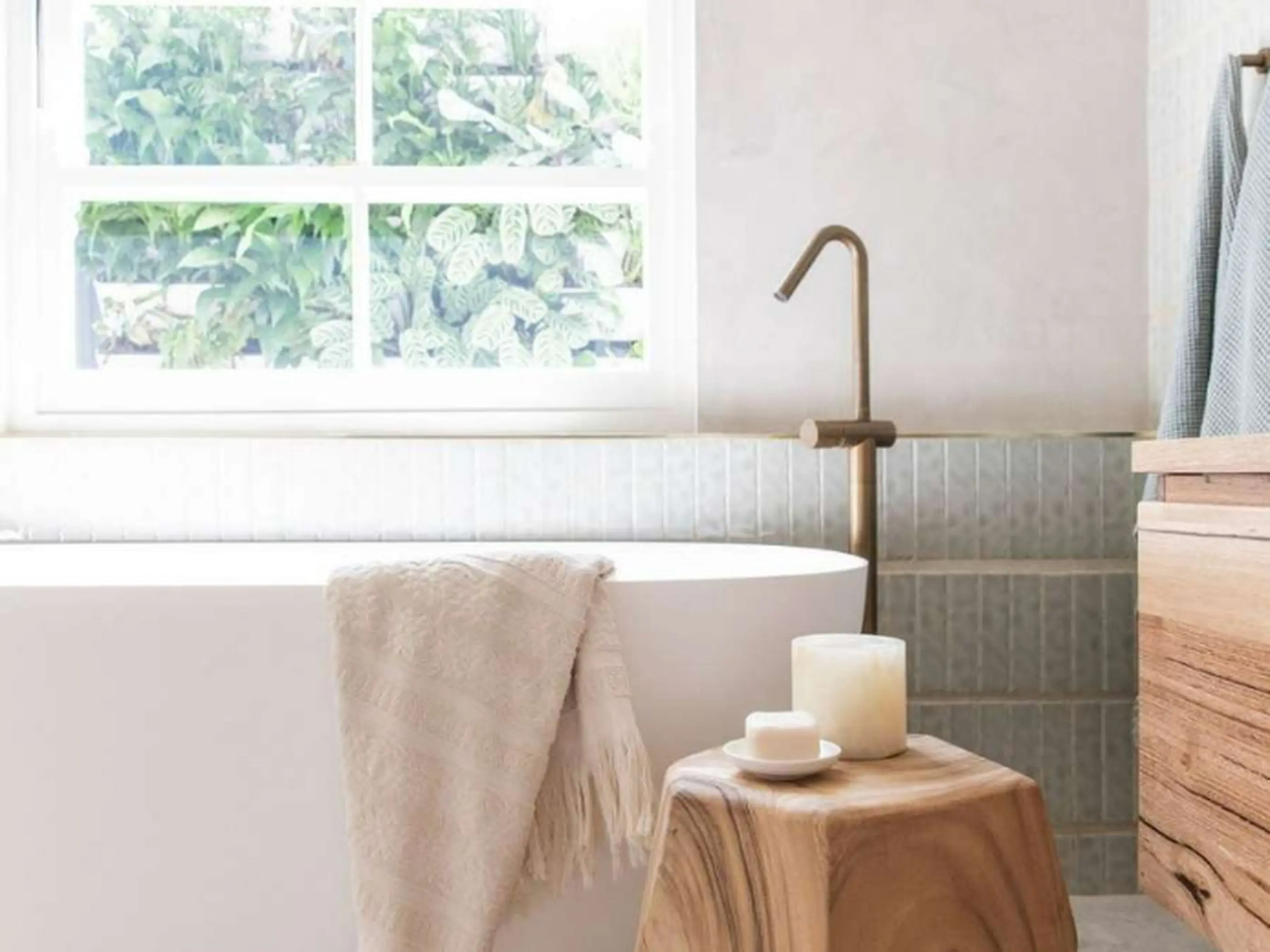 Modern bathroom with white tub, brass faucet, wooden stool with candles, and window overlooking greenery.