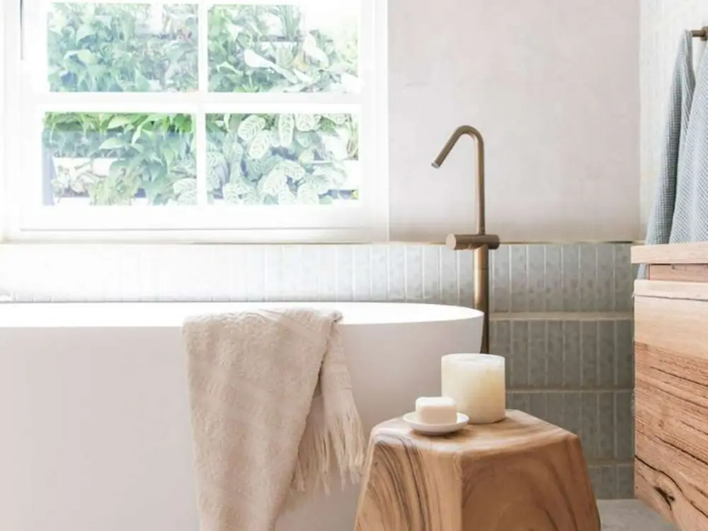 Modern bathroom with white tub, brass faucet, wooden stool with candles, and window overlooking greenery.