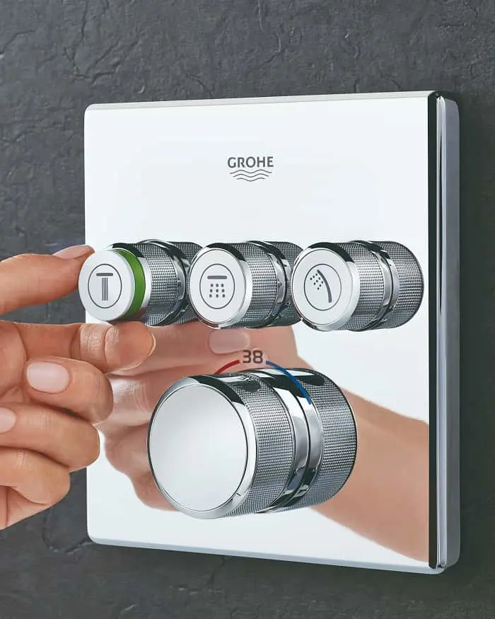 Finger pressing a button on a GROHE shower control panel with multiple chrome dials mounted on a gray wall.