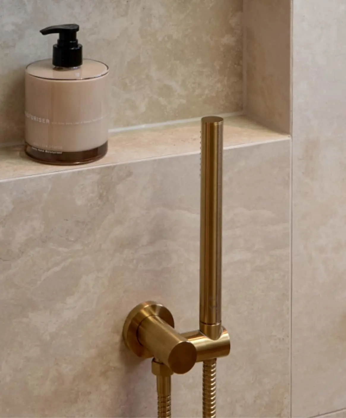 Beige pump bottle on marble shower shelf with brass handheld shower fixture mounted below.