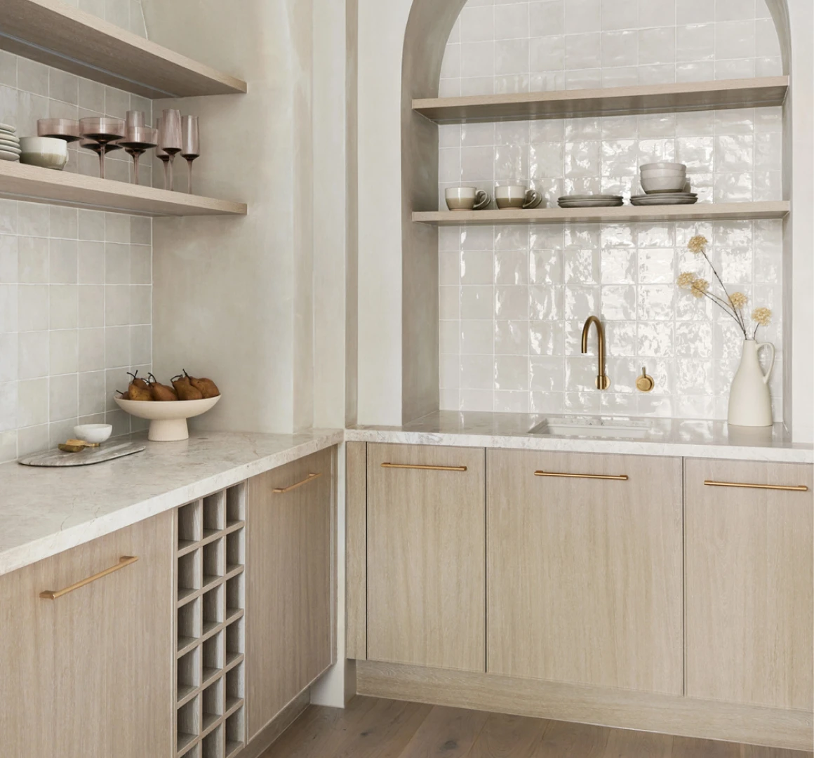 Modern kitchen with light wood cabinets, white tile backsplash, marble countertops, brass fixtures, and open shelving with dishware.