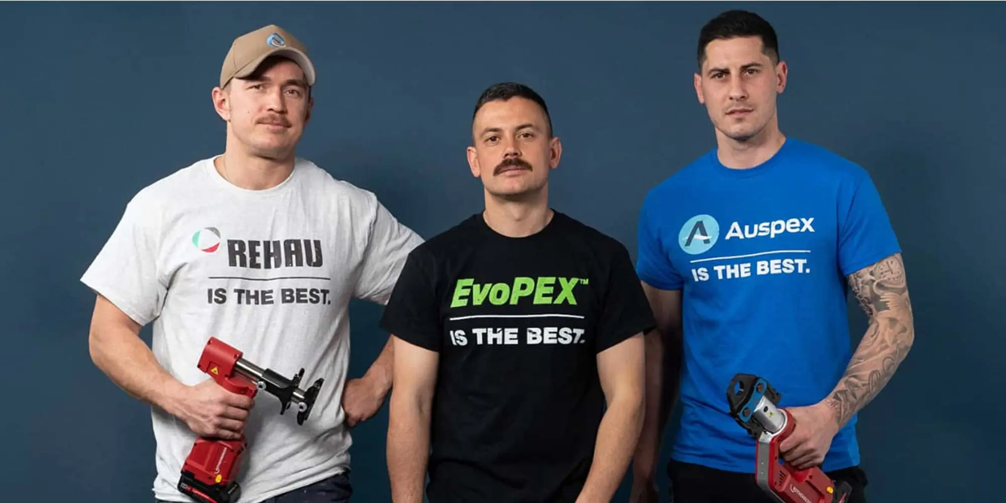Three men standing against blue background wearing branded t-shirts, two holding power tools.