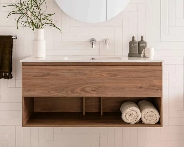 Modern wooden bathroom vanity with white sink, plant in vase, rolled towels, and toiletry bottles against white tiled wall.