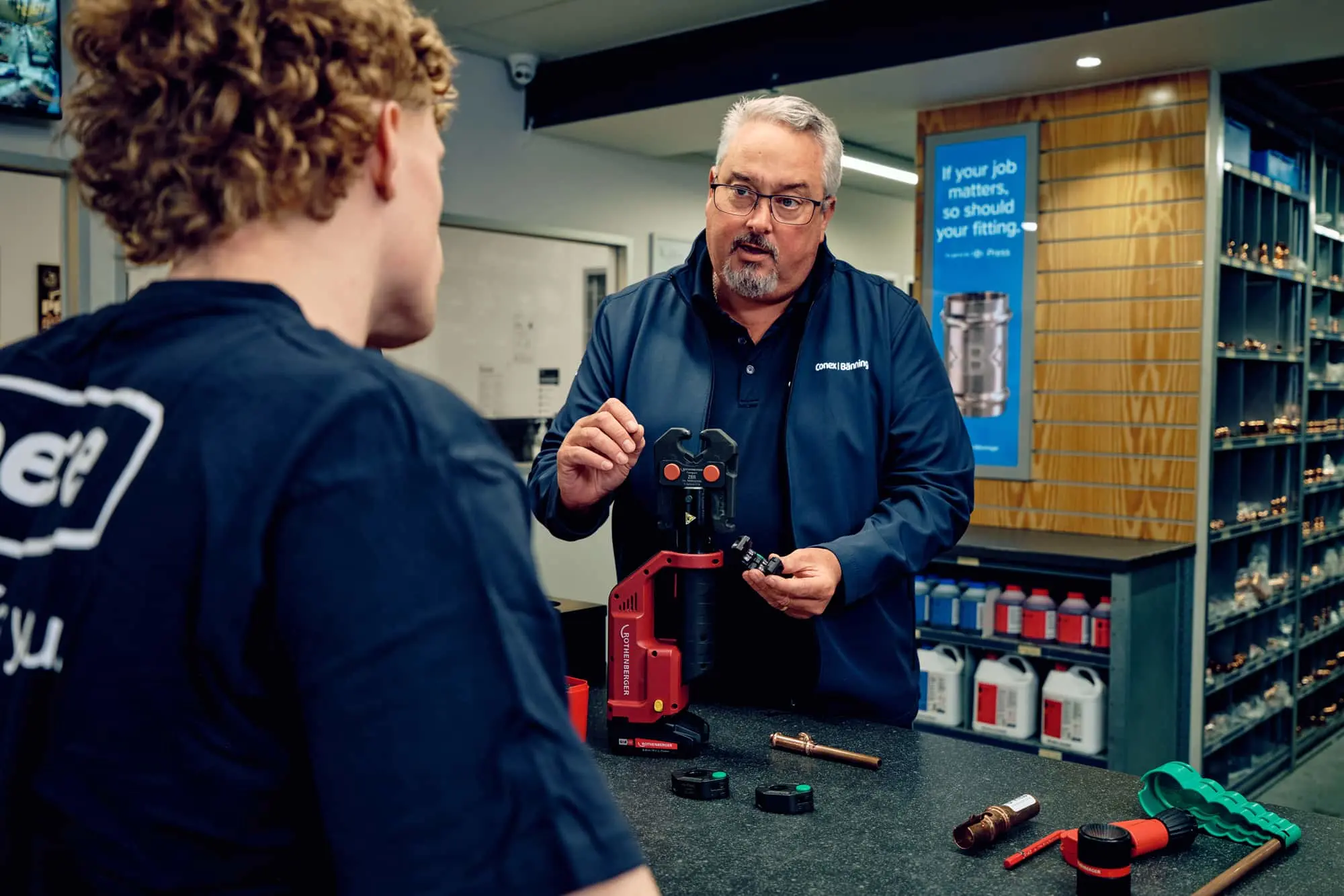 Instructor demonstrating plumbing equipment to a student at a workbench with tools and parts displayed.