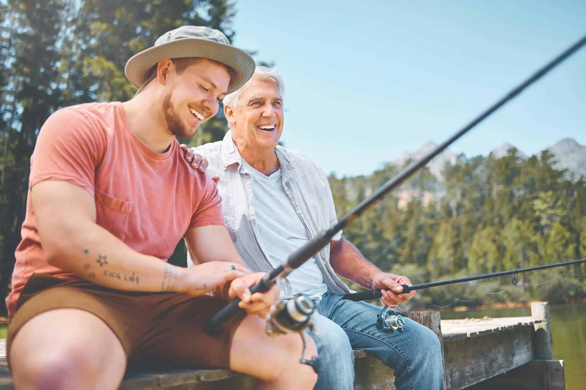 Two people fishing together on a wooden dock, smiling on a sunny day with forest in the background.