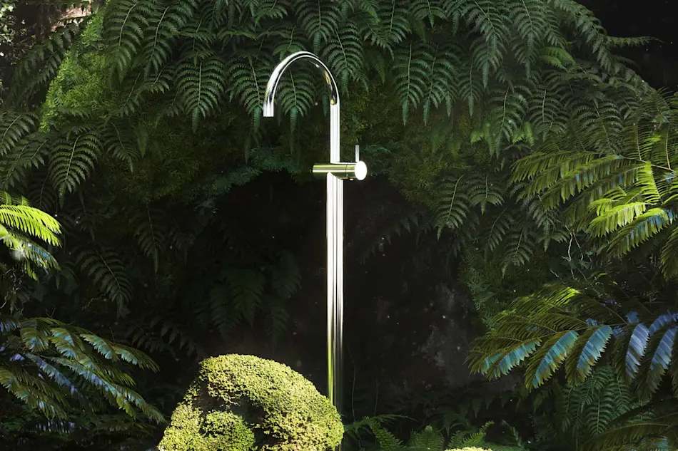 Modern chrome outdoor shower fixture surrounded by lush green ferns and a trimmed bush in a garden setting.