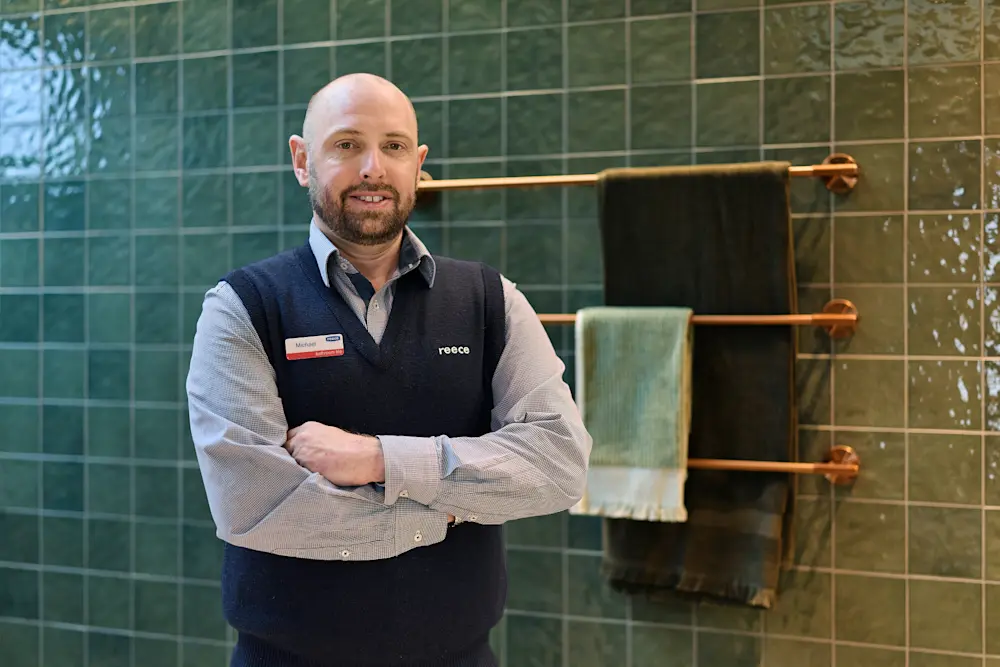 Person in navy vest and checkered shirt standing in front of green tiled wall with copper towel rails.