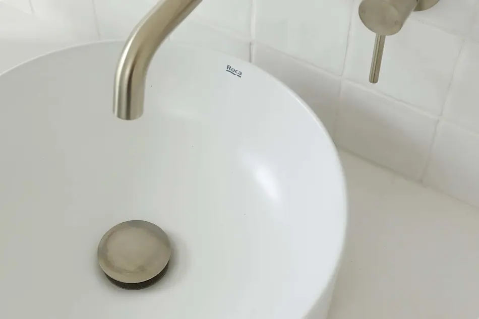 White Roca bathroom sink with stainless steel faucet and drain against white tiled wall.
