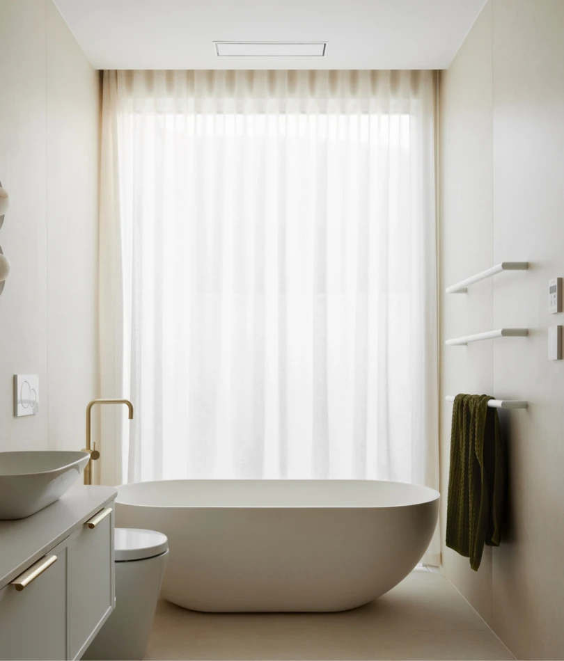 Modern white bathroom with freestanding tub, vessel sink, brass fixtures, and sheer curtains covering a large window.