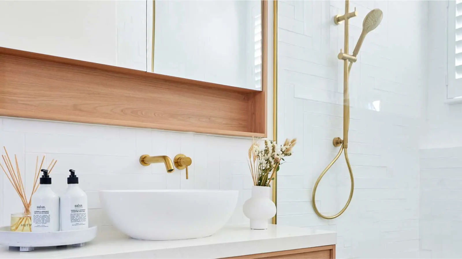 Modern bathroom with white vessel sink, brass fixtures, wooden accents, and gold shower system against white tile walls.