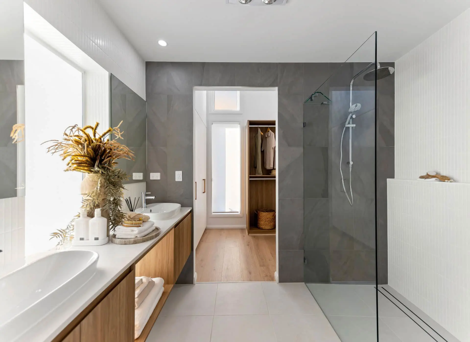 Modern bathroom with gray tile walls, glass shower, wooden vanity with double sinks, and walk-in closet visible through doorway.