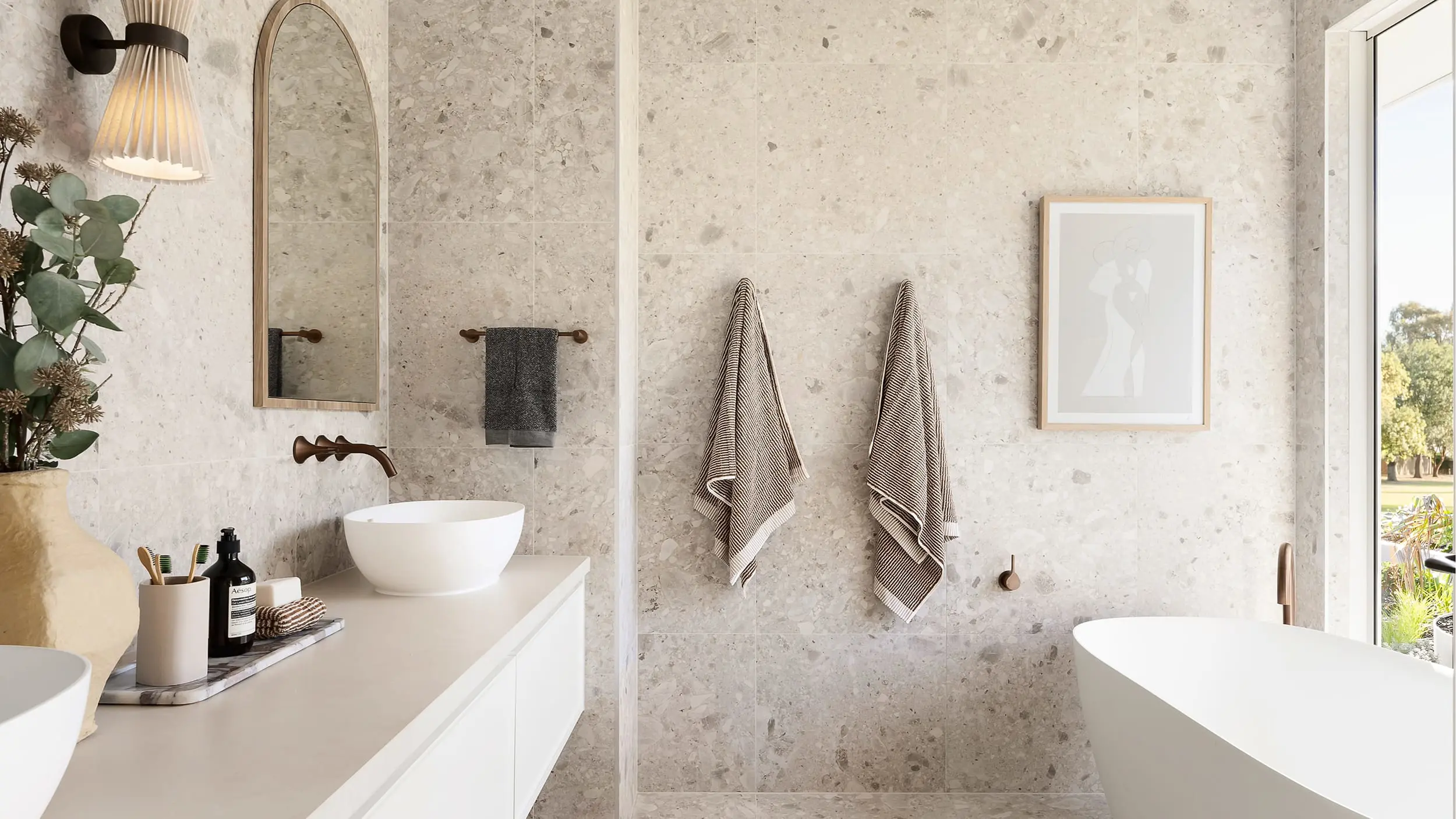 Modern bathroom with stone walls, white vanity, vessel sink, arched mirror, hanging towels, and bathtub with window view.