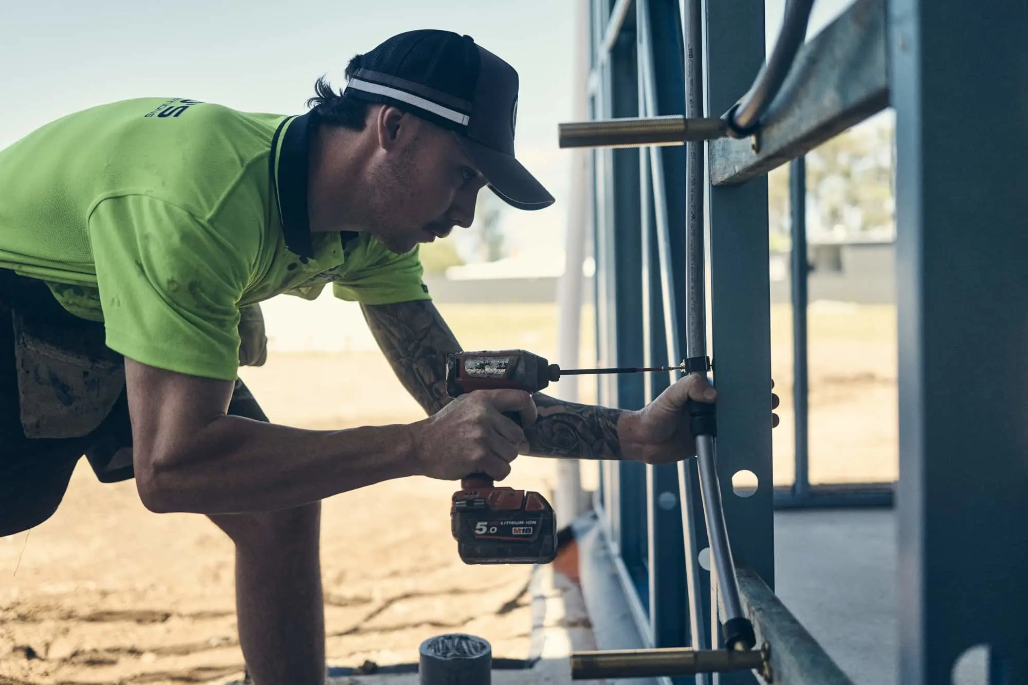 Construction worker in bright green shirt using power drill on metal framework at building site.