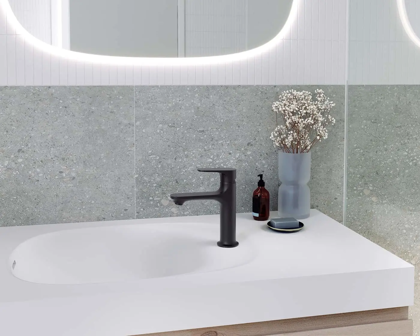 Modern bathroom sink with black faucet, gray terrazzo wall tiles, illuminated oval mirror, and decorative vase with flowers.