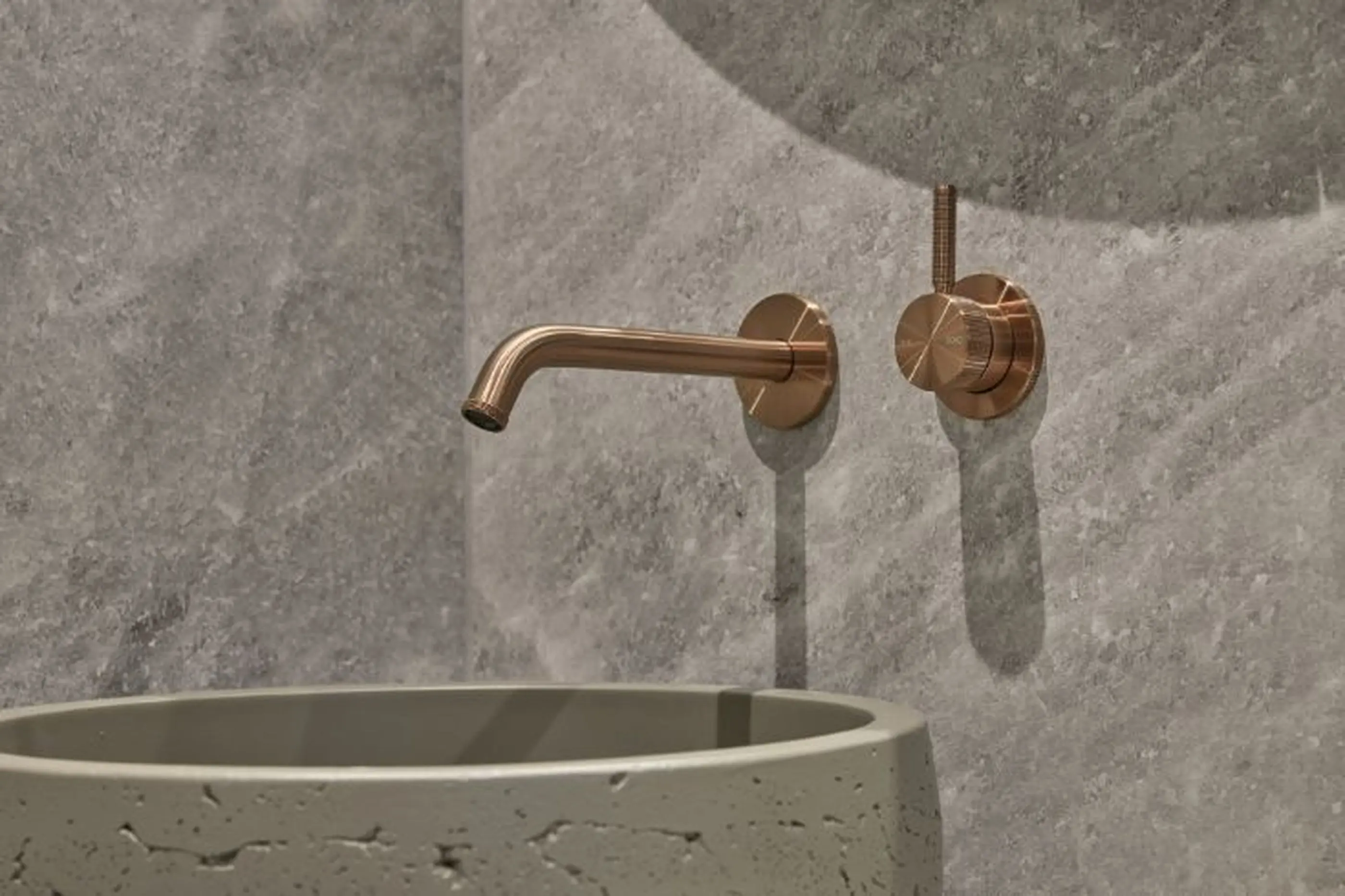 Copper wall-mounted faucet and control knob above a concrete basin in a bathroom with gray stone walls.
