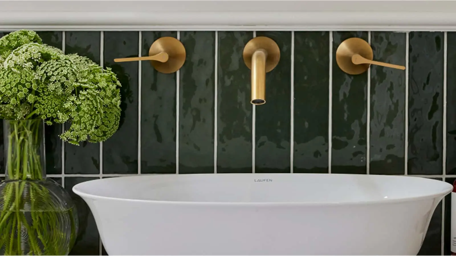 Modern bathroom with white sink, brass wall-mounted faucet, dark green tile backsplash, and fresh green plants.