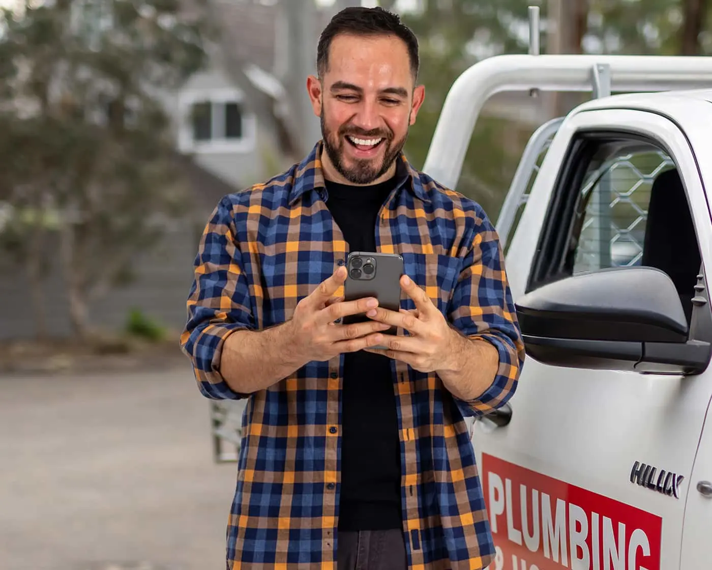 Person in blue and orange plaid shirt smiling while using smartphone next to a white plumbing service truck.