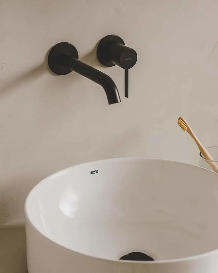 Modern bathroom sink with white Roca basin and matte black wall-mounted faucet, wooden toothbrush visible at edge.