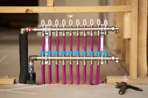 Radiant heating manifold with pink PEX pipes, flow meters, and valves mounted on wooden framing in construction setting.