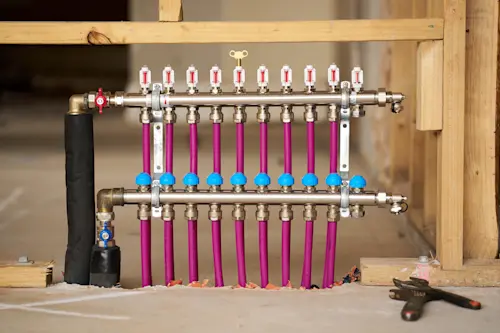 Radiant heating manifold with pink PEX pipes, flow meters, and valves mounted on wooden framing in construction setting.