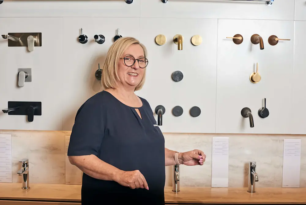 Woman in navy standing in bathroom showroom with various faucets and fixtures displayed on white wall behind her.