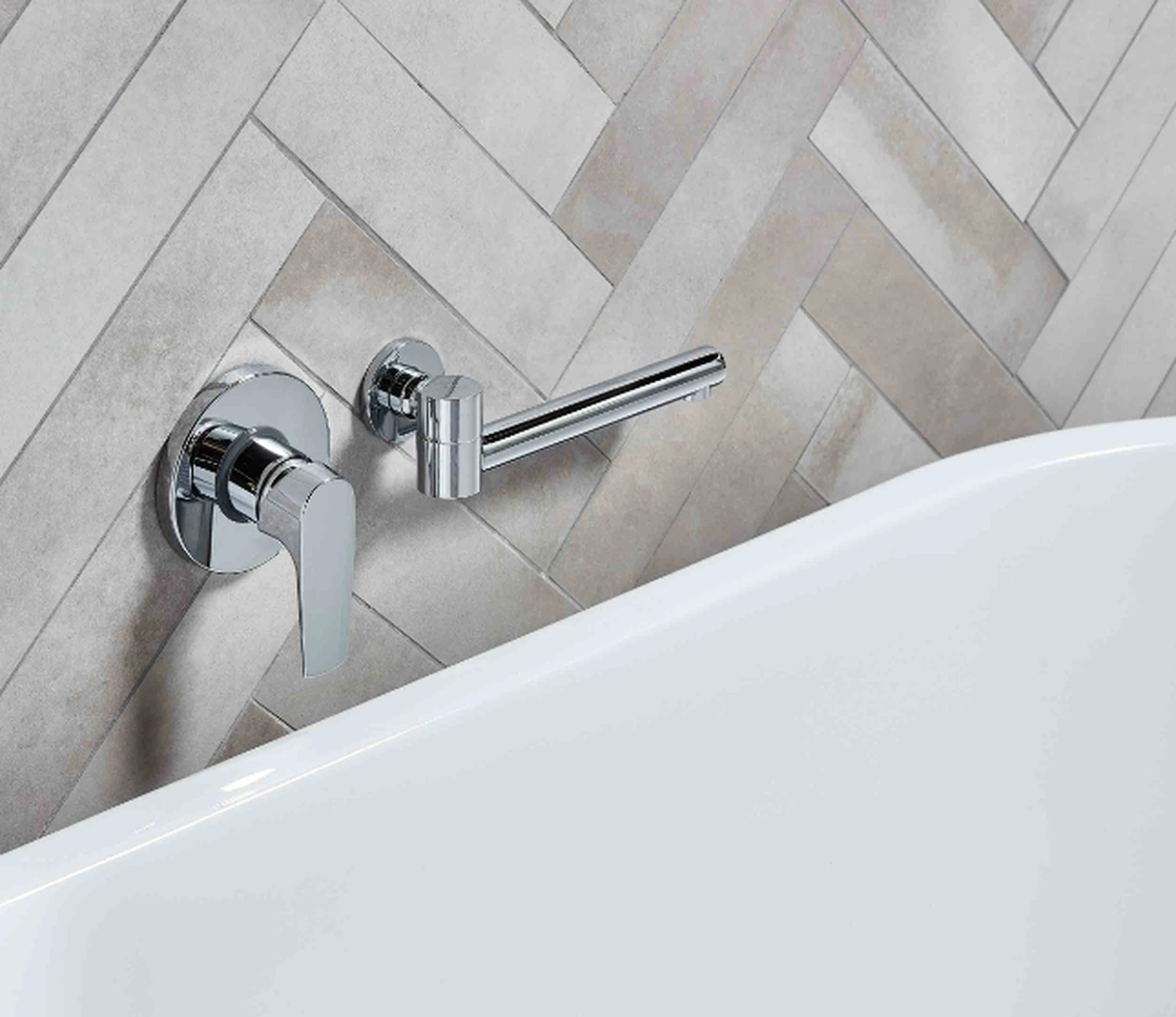 Modern chrome wall-mounted bathroom faucet with lever handle against light herringbone tile pattern, beside white bathtub.