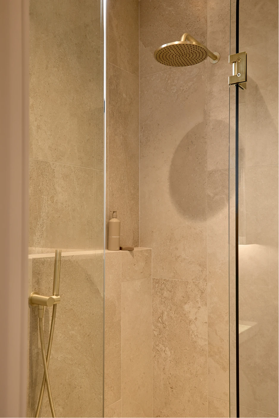 Beige tiled shower with gold rainfall showerhead, glass door, and built-in shelf with product bottle.