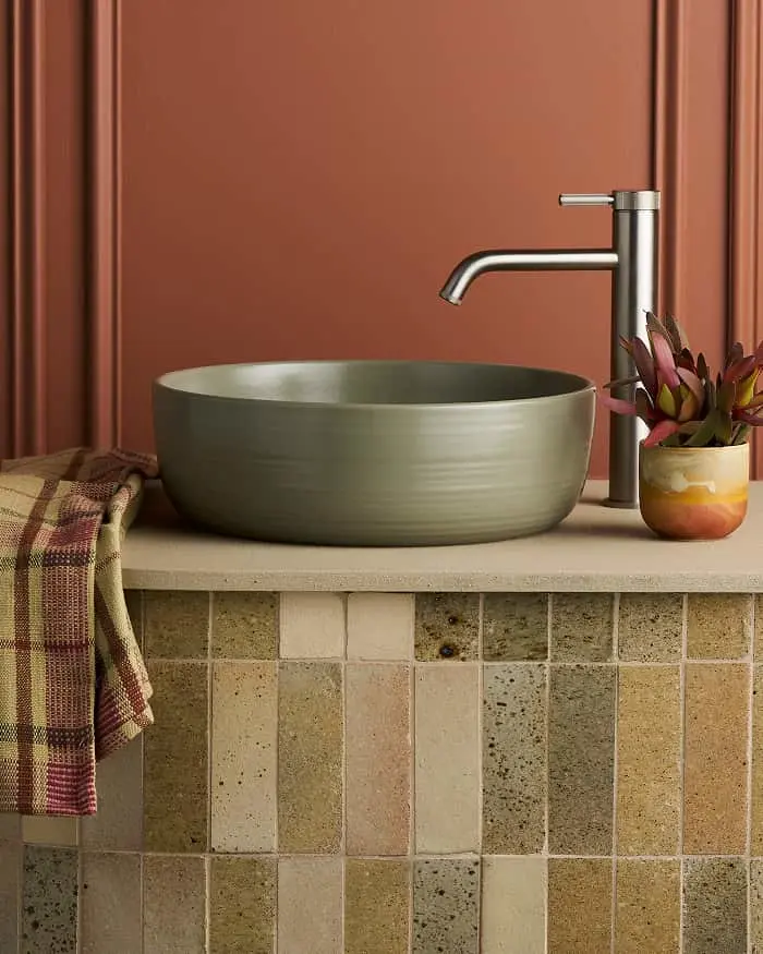 Green ceramic vessel sink on a stone tile vanity with a brushed nickel faucet, small plant, and plaid towel against a terracotta wall.