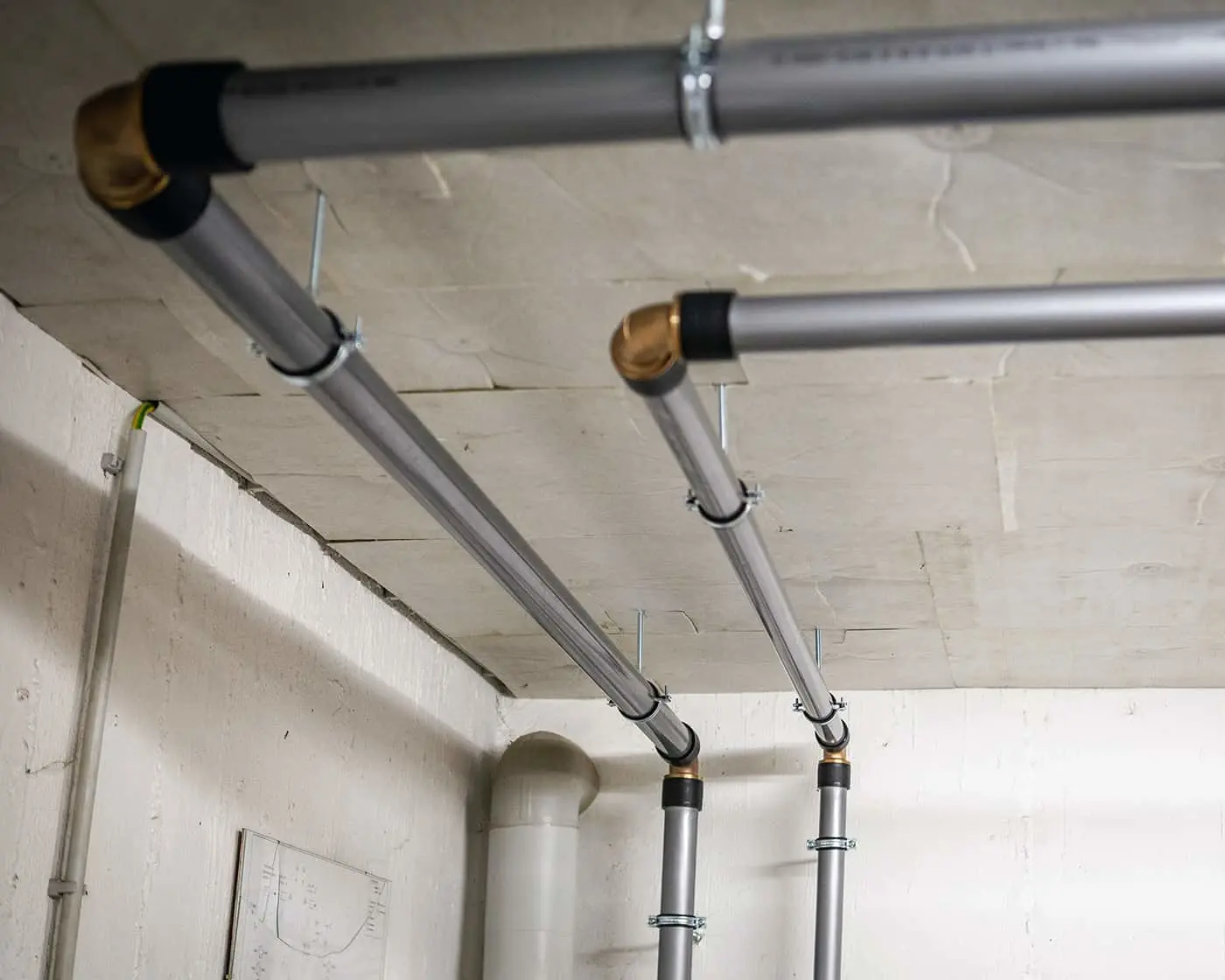 Metal plumbing pipes with brass and black fittings mounted to a concrete ceiling in a basement or utility room.