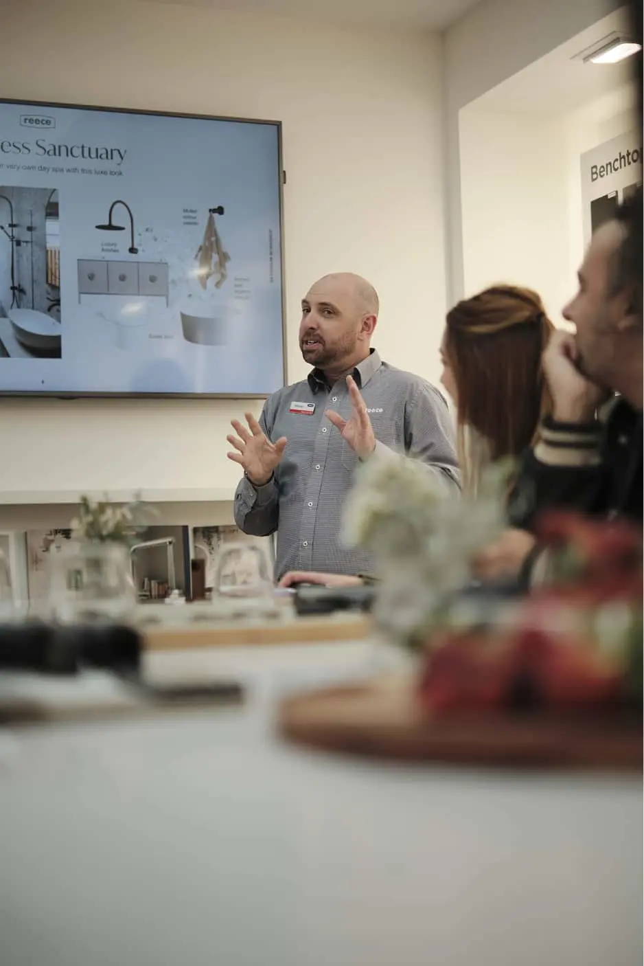 Staff member in gray shirt presenting bathroom design features on a display screen to customers in a showroom.