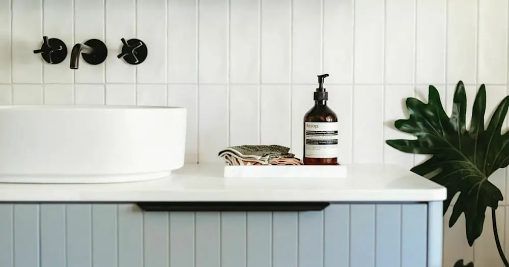 Modern bathroom sink with black wall fixtures, Aesop soap dispenser, folded washcloths, and green plant on white countertop.