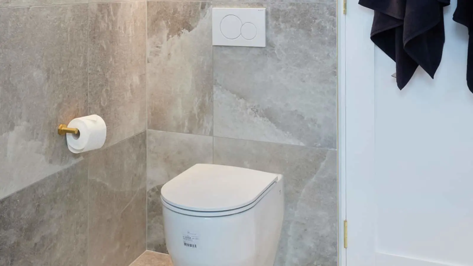 Modern bathroom with white toilet, gray stone tile walls, gold toilet paper holder, and black towel hanging nearby.
