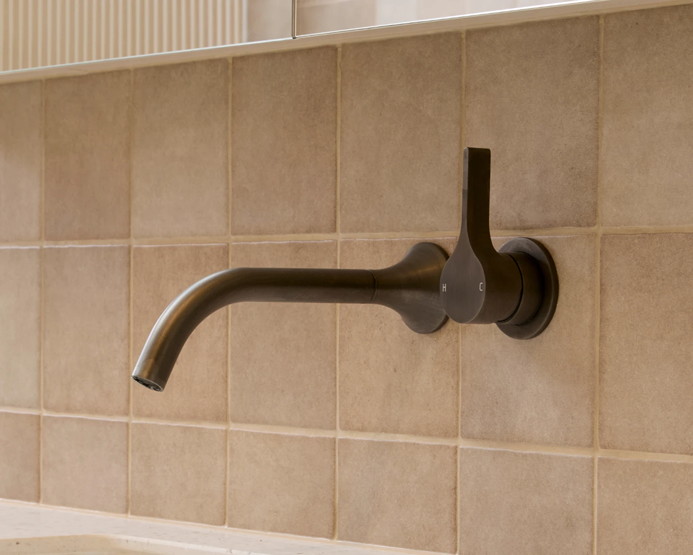 Modern dark bronze wall-mounted bathroom faucet with curved spout against beige tiled wall.