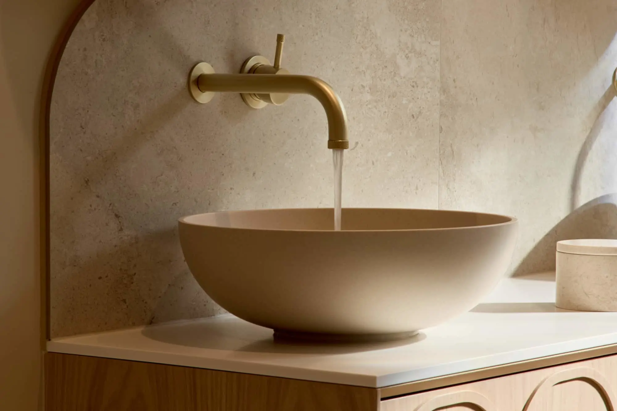 A round beige vessel sink on a light wood vanity with a gold wall-mounted faucet running water, set against a stone tile wall.