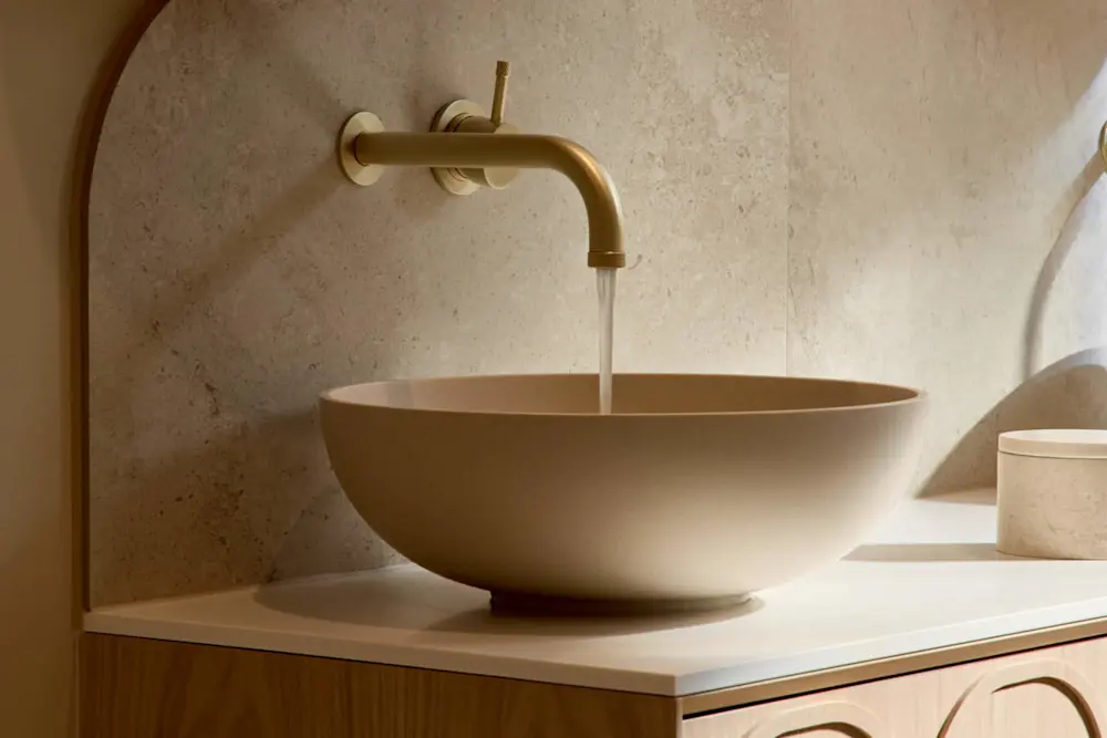 A round beige vessel sink on a light wood vanity with a gold wall-mounted faucet running water, set against a stone tile wall.