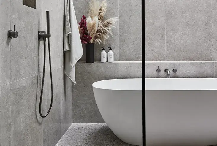 Modern gray bathroom with white freestanding tub, glass shower partition, and dried flower arrangement on shelf.