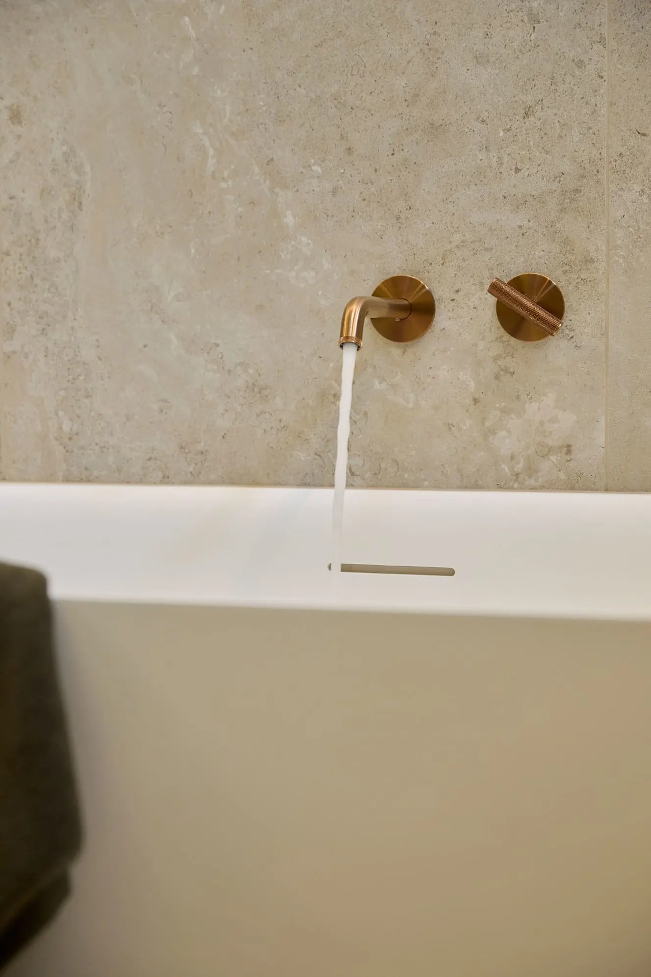 Modern bathroom with white bathtub and brass wall-mounted faucet running water against beige stone wall.
