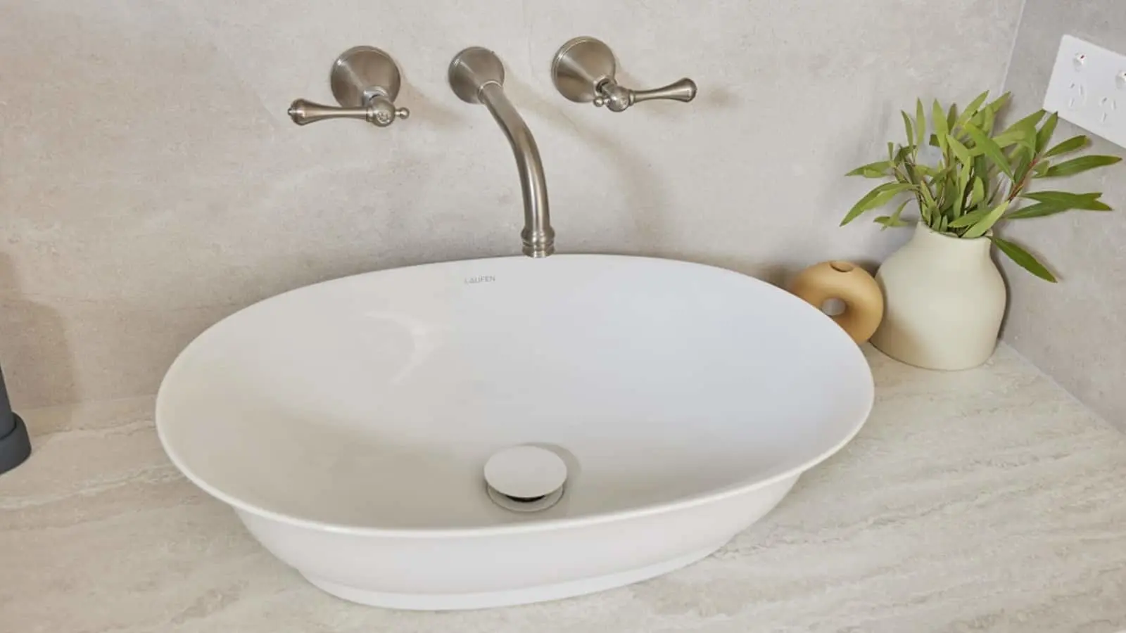 White oval bathroom sink with brushed nickel wall-mounted faucet, beige countertop, and small plant in cream vase.