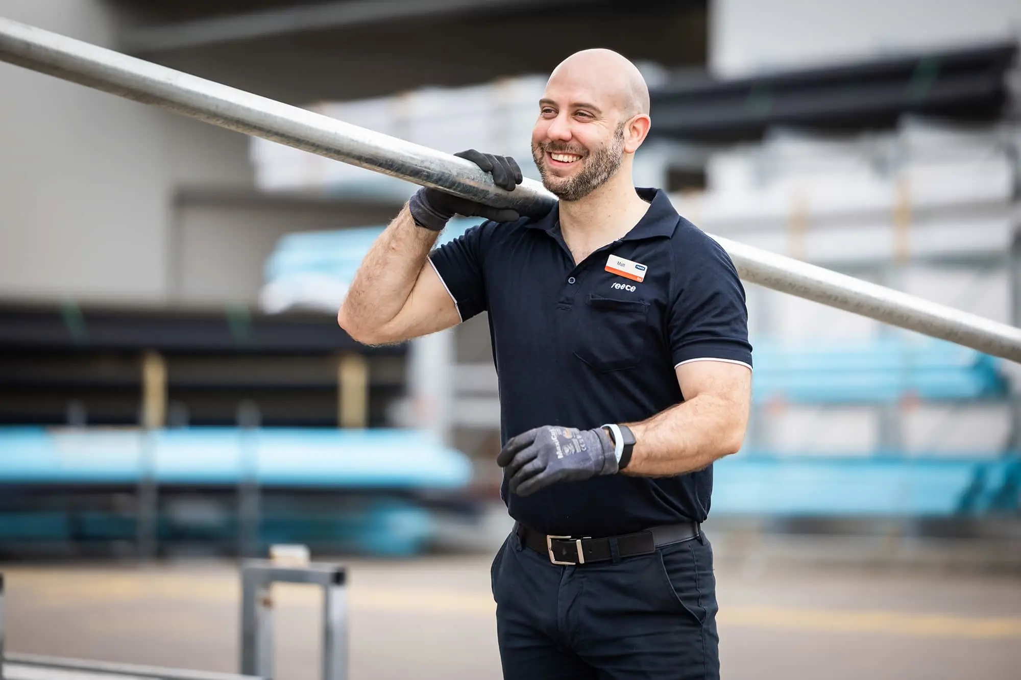 Worker in navy uniform and gloves smiling while carrying a metal pole at a construction site.