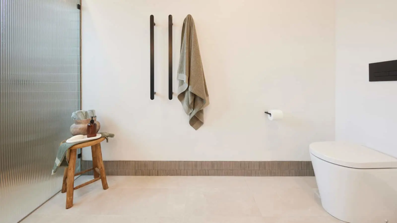 Minimalist bathroom with white walls, towel hanging on black hooks, wooden stool with toiletries, and white toilet.