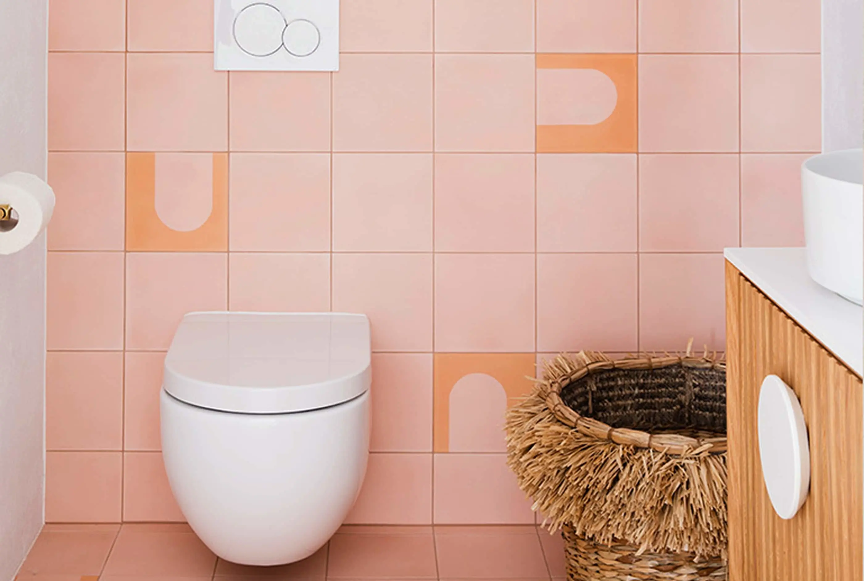 Modern bathroom with white toilet, pink tiled walls with orange accents, woven basket, and wooden vanity.