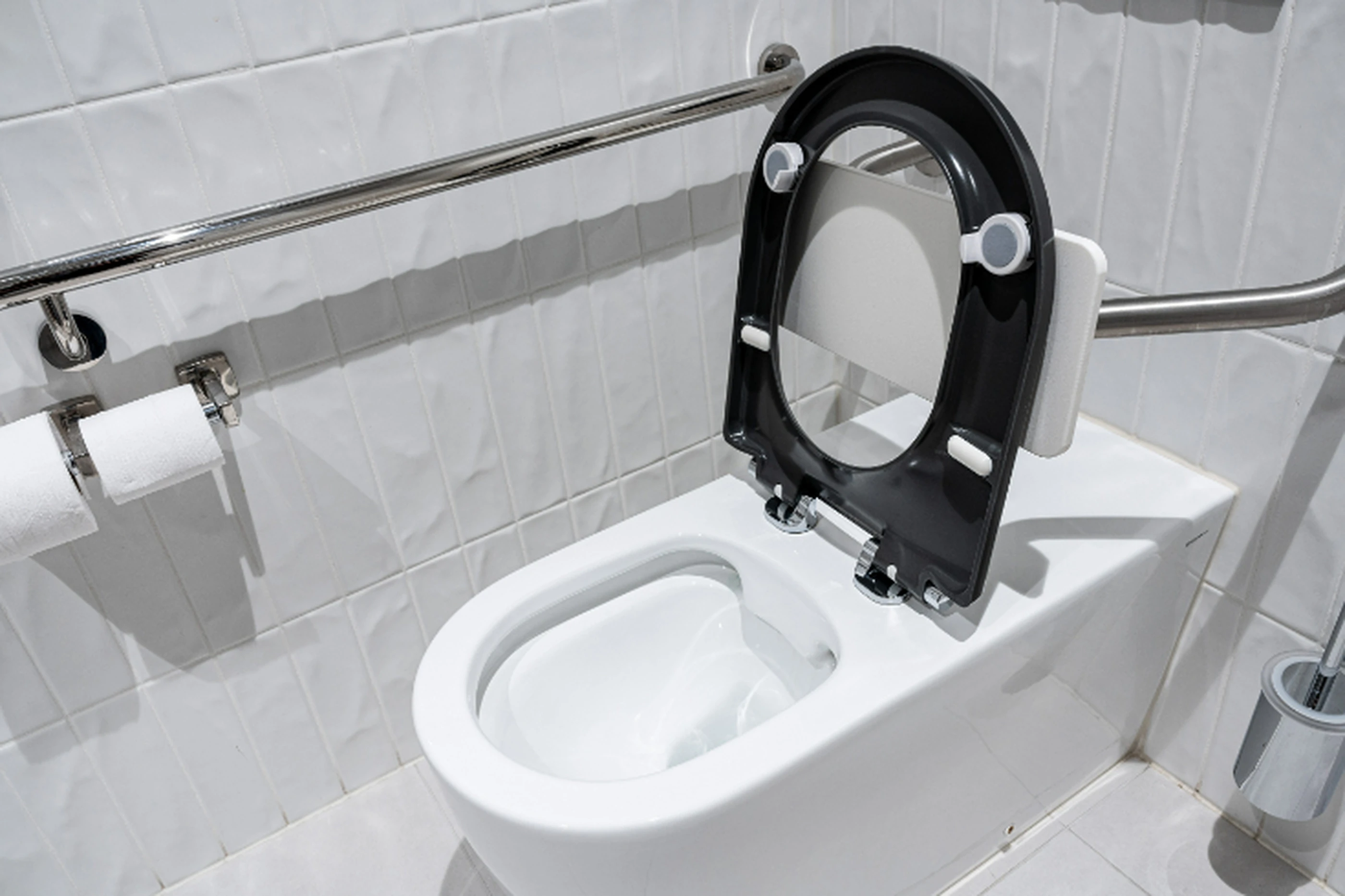 White toilet with black seat lid raised, metal grab bars on tiled wall, and toilet paper holder visible.