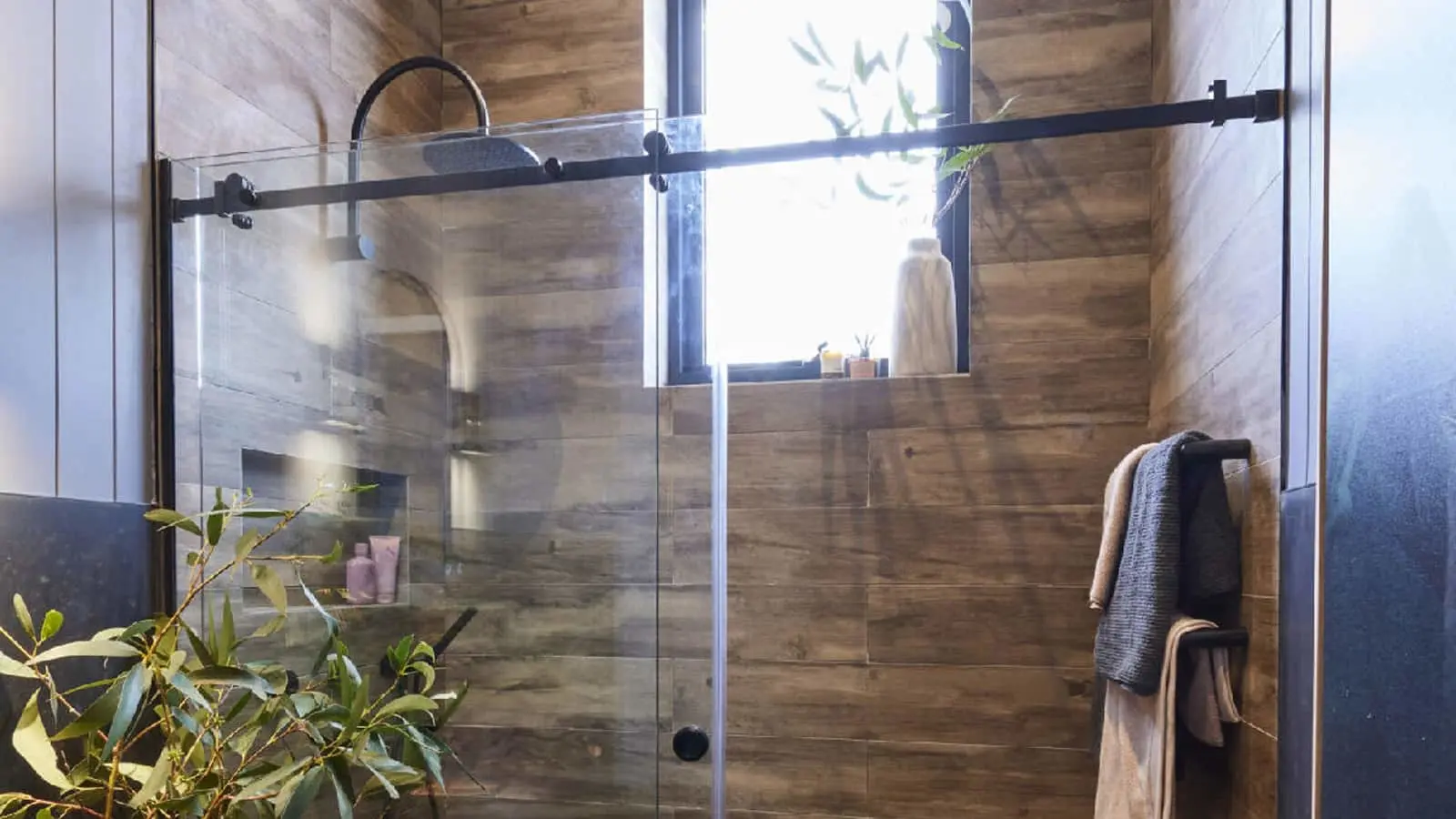 Modern bathroom with wood-look tile walls, glass shower door, rainfall showerhead, window, and hanging towels.