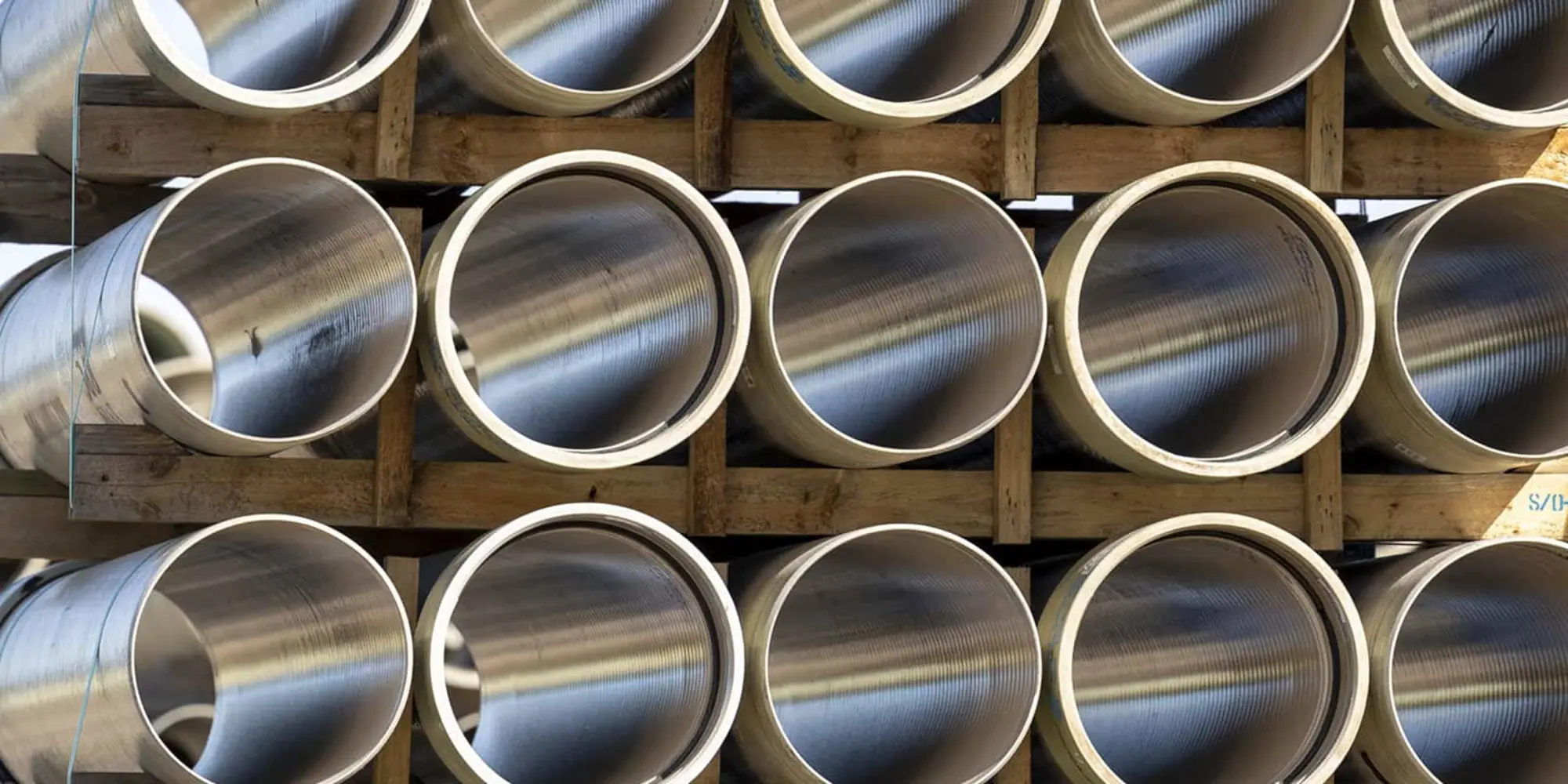 Stacked metal pipes with threaded ends arranged in rows on wooden supports in a storage or construction area.