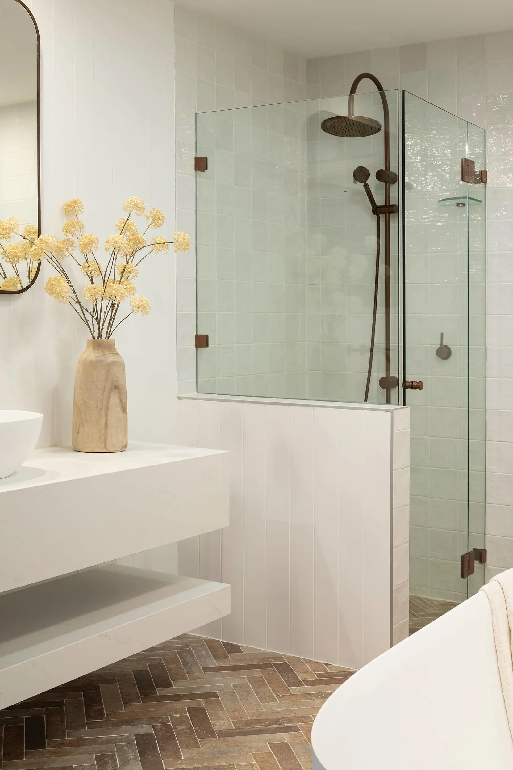 Modern bathroom with glass shower, bronze fixtures, white tiled walls, wooden vase with yellow flowers, and herringbone floor.