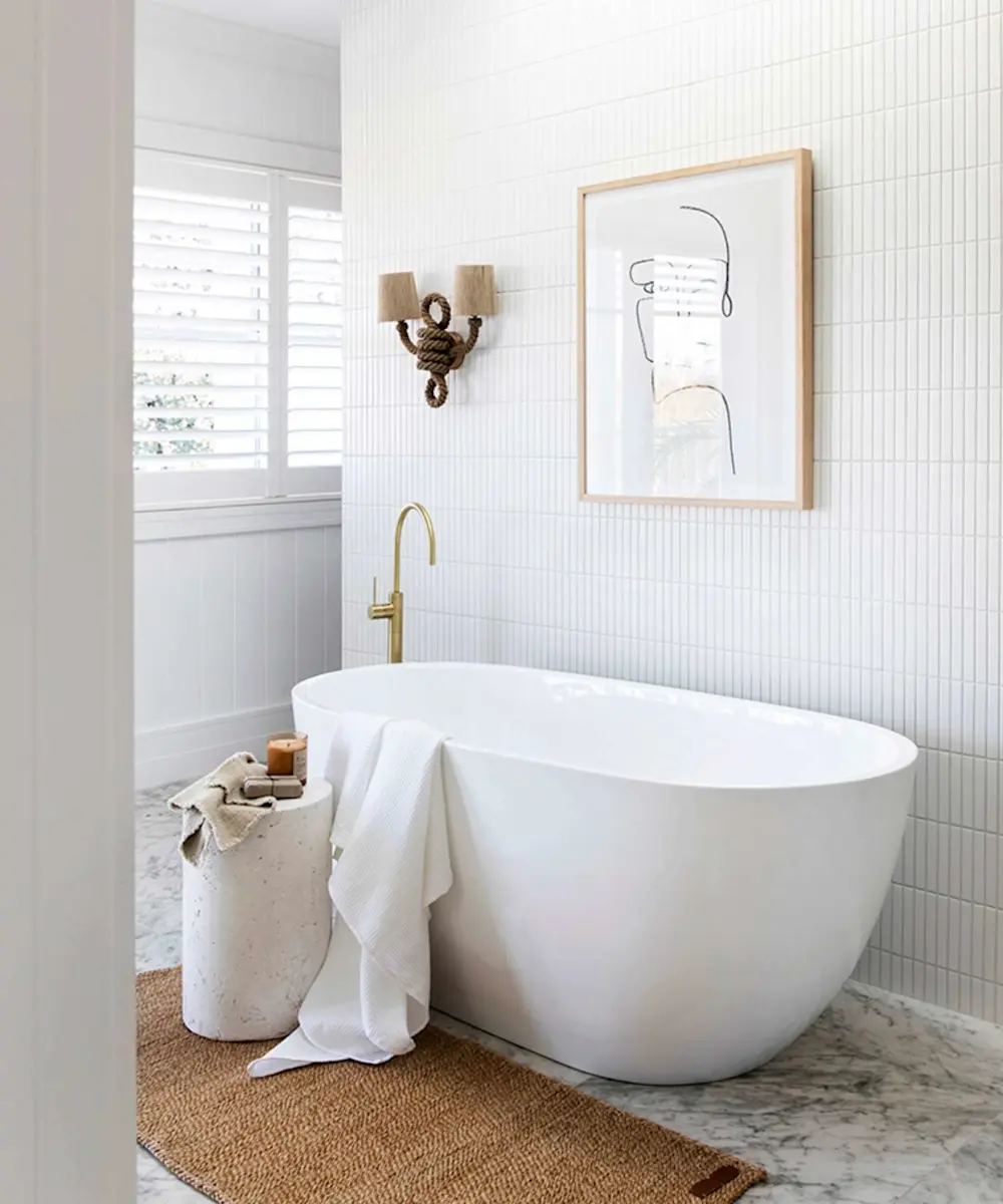 Modern white bathroom with freestanding tub, rope wall sconce, minimalist artwork, and jute rug on marble floor.