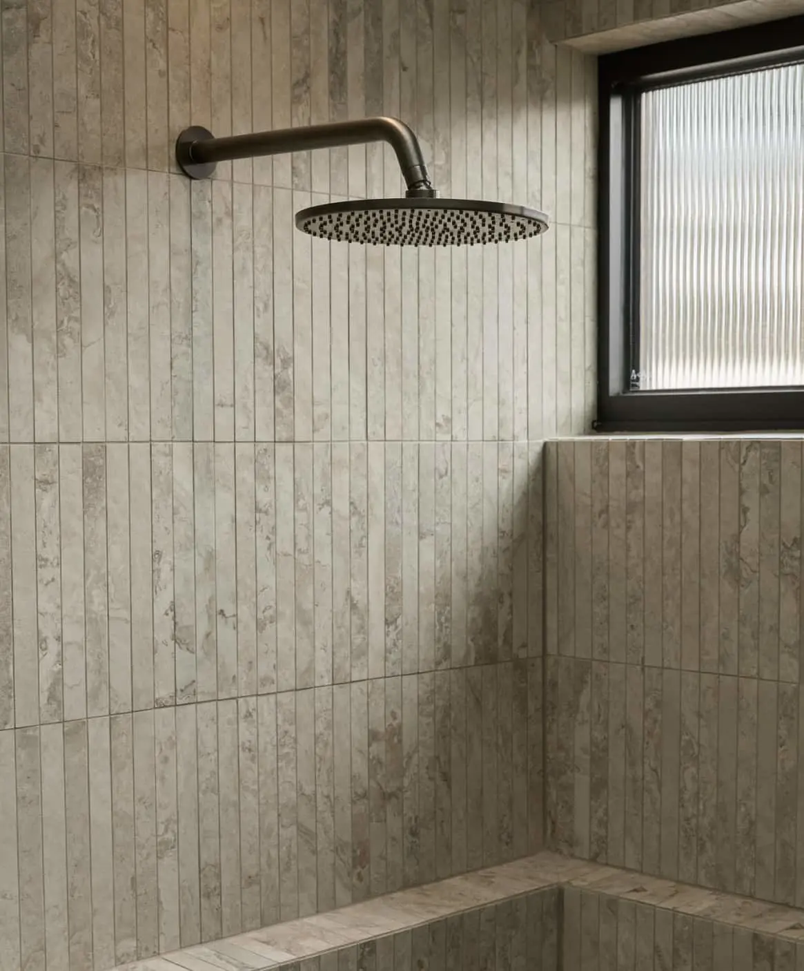 Modern shower with round gunmetal rainfall showerhead mounted on beige tiled walls next to a window.