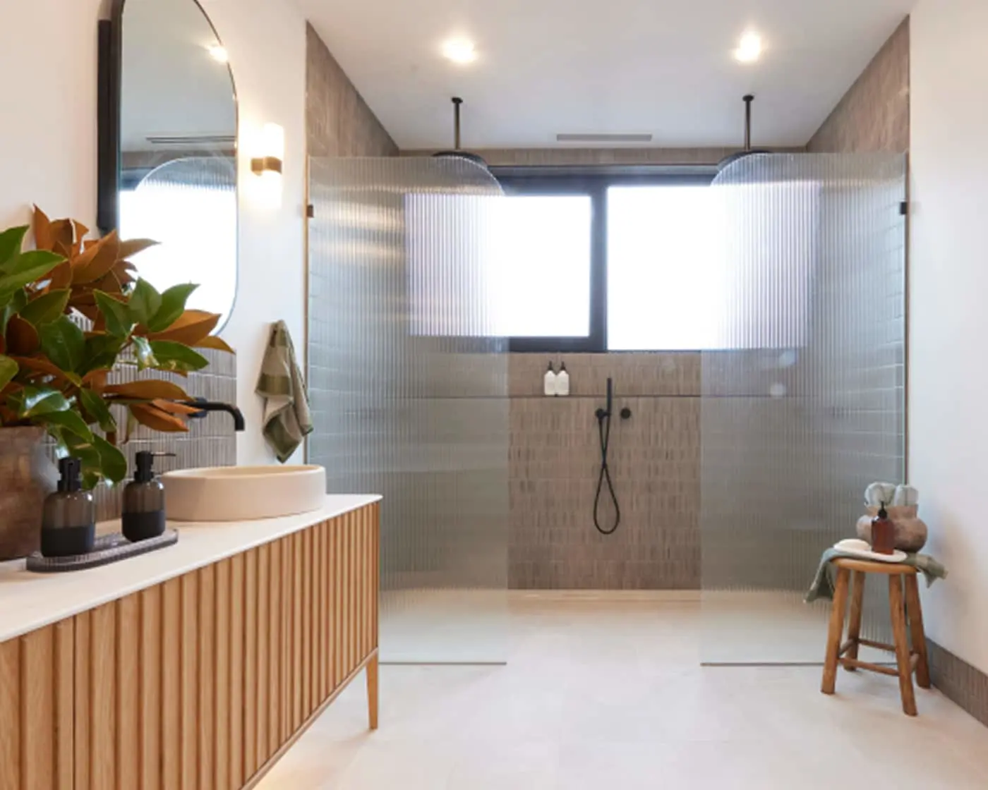 Modern bathroom with wooden vanity, glass shower enclosure, potted plant, and wooden stool with toiletries.